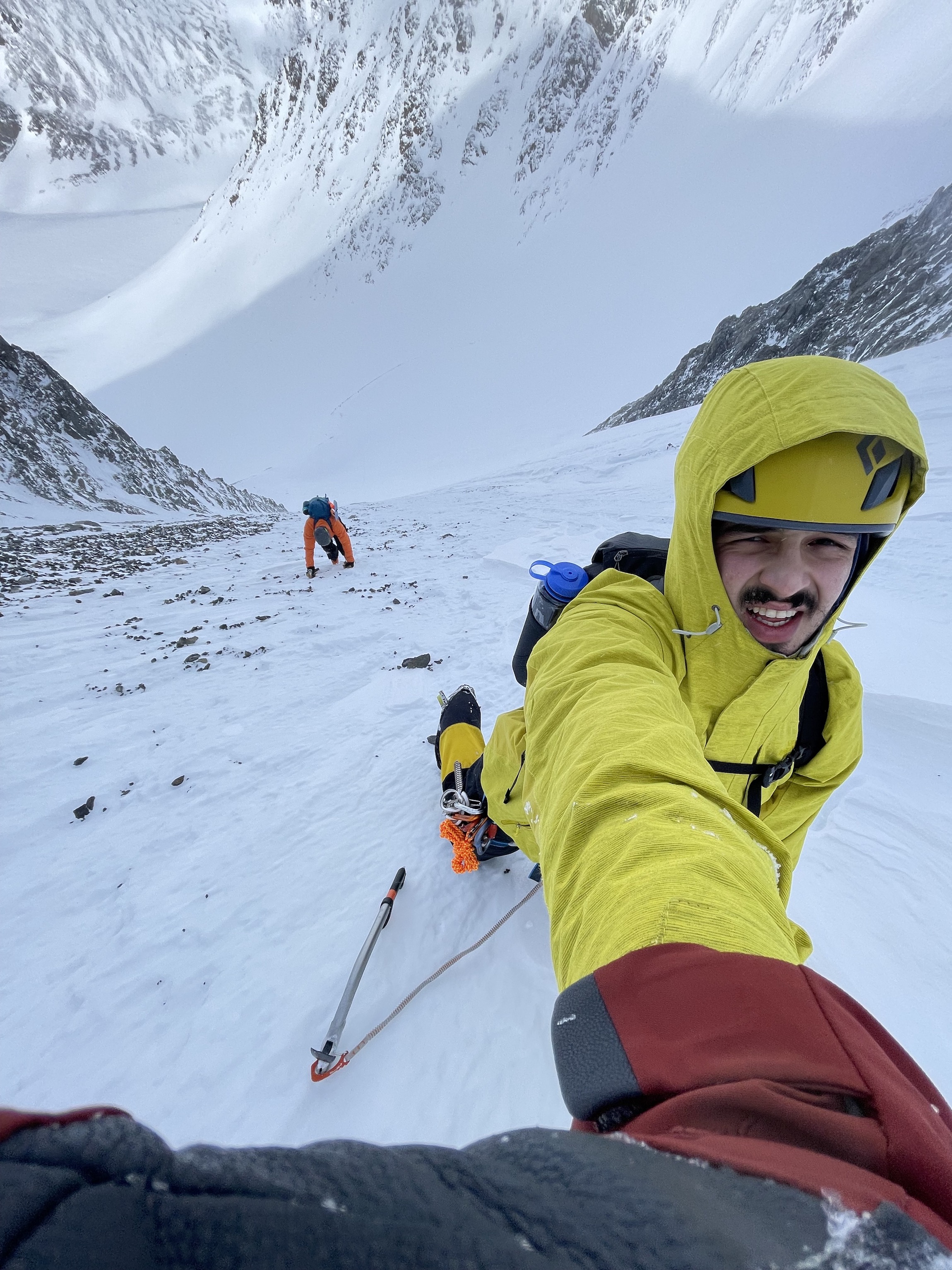 close-up of a mountain climber in a yellow jacket jacket and helmet reaching up towards the camera with another climber lower down the slope